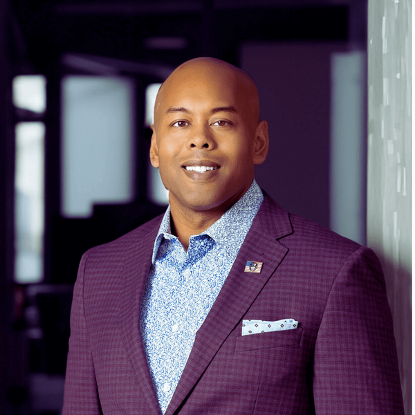Man wearing a purple checkered suit with a blue shirt and patterned pocket square, standing indoors.