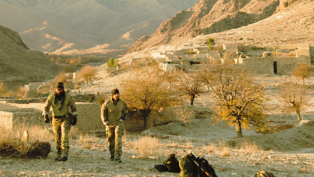 Two people in military gear walking through a desert landscape with mountains in the background.